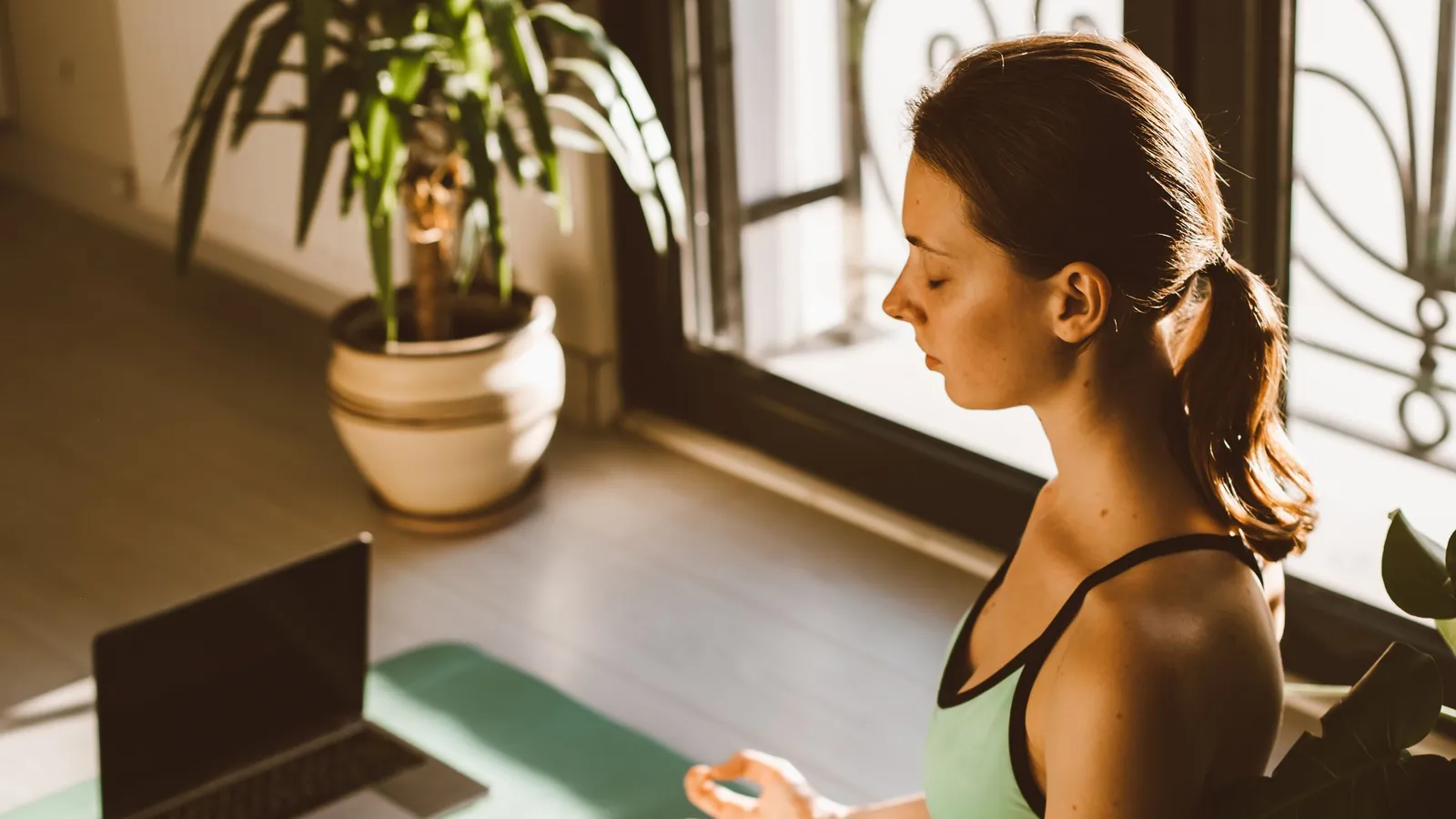 Eine Frau meditiert auf einer Yogamatte in einem sonnigen Raum, neben ihr ein aufgeklappter Laptop